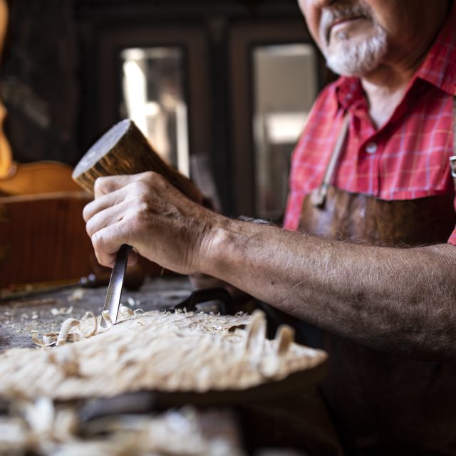 An old fashioned experienced senior carpenter holding knife and hammer carving wooden board in his woodworking workshop.